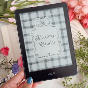 Op de afbeelding: Een zwarte Kindle e-reader met een aangepast scherm met een wit en grijs ruitpatroon en de tekst "Olivia's Kindle" in een decoratief kader. Het apparaat is omringd door bloemen.