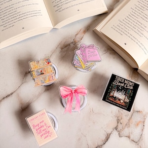 Könnte beinhalten: Eine Sammlung von Buch-Accessoires, darunter eine rosa Schleife, ein Stapel Bücher mit dem Text "Book Girls", eine Bibliothekskarte mit Text, ein Bücherregal und ein Design "Tired Moms Book Club". Die Artikel sind auf einer Marmoroberfläche angeordnet.