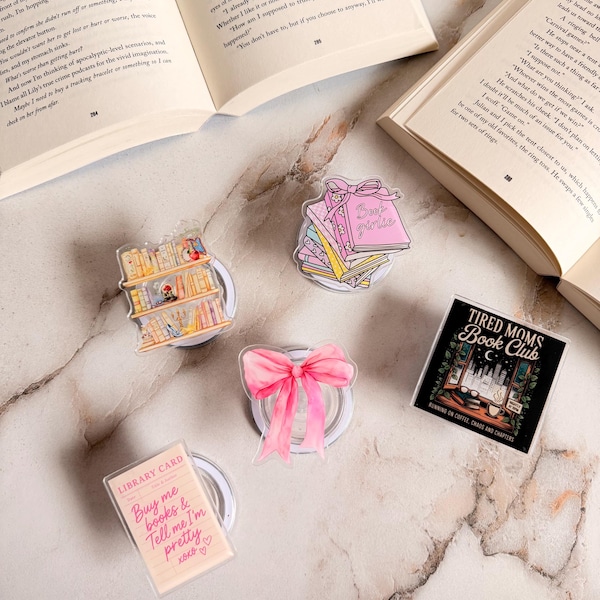 May include: A collection of decorative book-themed accessories, including a pink bow, a stack of books with "Book Girls" text, a library card with text, a bookshelf, and a "Tired Moms Book Club" design. The items are arranged on a marble surface.