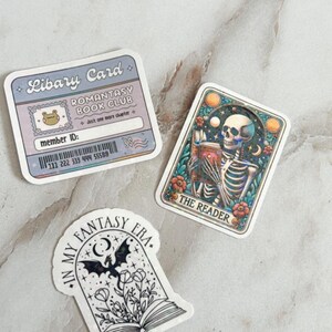 Op de afbeelding: Drie stickers met fantasythema's. De eerste sticker is een bibliotheekkaart met een kikkermascotte en de tekst "Romantasy Book Club". De tweede sticker toont een skelet dat een boek leest met de tekst "The Reader". De derde sticker toont een boek met een draak die erboven vliegt en de tekst "In My Fantasy Era."