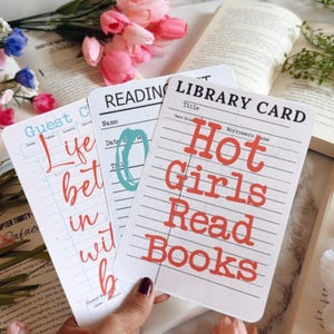 May include: Three white library cards with red and blue text, including "Hot Girls Read Books." The cards are held near open books and pink flowers, suggesting a reading theme.