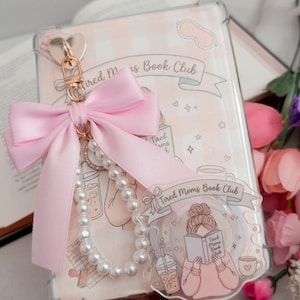 May include: A clear phone case with a pink bow, pearl beads, and a gold heart-shaped clasp. The case features the text "Tired Moms Book Club" and a cartoon illustration of a woman reading. The case is decorated with hearts and stars.