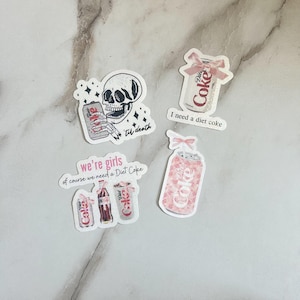 Könnte beinhalten: Eine Sammlung von Aufklebern mit Diet Coke-Dosen und -Flaschen. Ein Aufkleber zeigt einen Totenkopf, der eine Dose mit dem Text "'til death" hält. Weitere Aufkleber enthalten die Sätze "I need a diet coke" und "we're girls of course we need a Diet Coke".