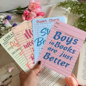 May include: A hand holds four colorful, lined paper cards with text. The cards are pink, blue, peach, and light green. The pink card reads "Boys in Books are just Better." Other cards say "Guest Check" and "Library".