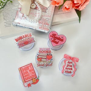 Könnte beinhalten: Eine Sammlung klarer Handyhüllen und PopSockets mit Buchmotiven. Die Hüllen haben eine Schleife und ein Perlenarmband. Die PopSockets zeigen Sätze wie "Romance Reader" und "Hot Girl Book Club" in Rosa- und Rottönen.