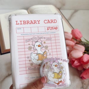 May include: A clear phone case with a pink and white library card design. The card features a cartoon goose reading a book, the text "literally just a girl", and the words "avoiding reading". A matching sticker is also visible.