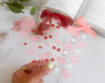 Può includere: Tre segnalibri in plastica trasparente con disegni di ciliegie e fiocchi rosa e bianchi. I segnalibri sono sovrapposti.