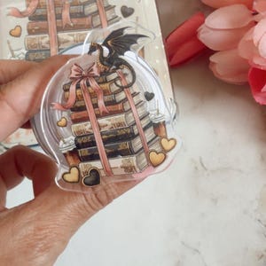 Könnte beinhalten: Ein klarer Acryl-Charm mit einem Stapel Bücher, die mit einem rosa Band zusammengebunden sind, gekrönt von einem schwarzen Drachen. Goldene und schwarze Herzen umgeben die Bücher. Ein passendes Design ist im Hintergrund sichtbar.