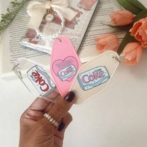 Könnte beinhalten: Drei Schlüsselanhänger im Vintage-Stil mit Diet Coke-Branding. Einer ist transparent mit rotem und weißem Text, ein anderer ist rosa mit einem Herzdesign und der dritte ist beige. Die Schlüsselanhänger werden in einer Hand mit einem Ring gehalten.