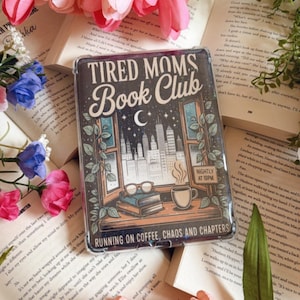 Op de afbeelding: Een telefoonhoesje met de tekst "TIRED MOMS Book Club" en "RUNNING ON COFFEE, CHAOS AND CHAPTERS". De hoes heeft een skyline, een halve maan en een kopje koffie. Roze en perzikkleurige tulpen en open boeken omringen de hoes.
