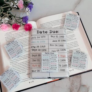 May include: A clear phone case with the words "Date Due" printed on it, along with a list of dates. Several small cards with the same text and dates are scattered around an open book, with a floral arrangement in the background.
