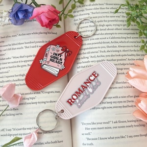 May include: Two motel-style keychains. One is red with "HOT GIRLS READ!" text and a book graphic. The other is pink with "ROMANCE Book Club" text. Both have silver key rings. Flowers and a book are in the background.