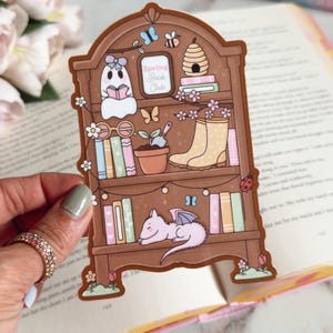 May include: A brown sticker featuring a bookshelf design with books, a ghost reading, a beehive, and a sleeping dragon. The sticker has the text "Spring Book Club" and is decorated with flowers and butterflies.