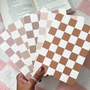 Può includere: Cinque carte a scacchi con diverse tonalità di marrone e rosa. Le carte sono tenute in una mano con un braccialetto d'oro al polso.