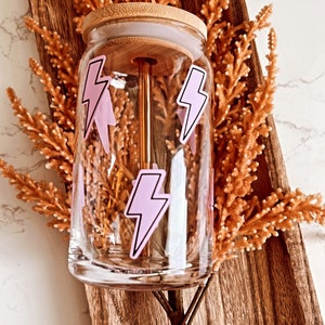 May include: A clear glass tumbler with a bamboo lid and a pink lightning bolt design. The tumbler is sitting in a wooden tray with dried flowers.