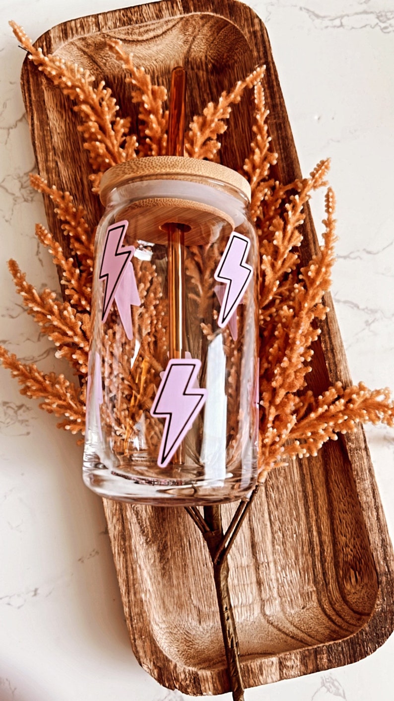 May include: A clear glass tumbler with a bamboo lid and a pink lightning bolt design. The tumbler is sitting in a wooden tray with dried flowers.
