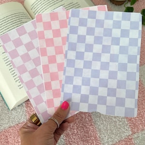 Peut inclure: Trois feuilles de papier à carreaux en rose, violet clair et blanc. Les feuilles sont empilées les unes sur les autres et sont tenues par une main.