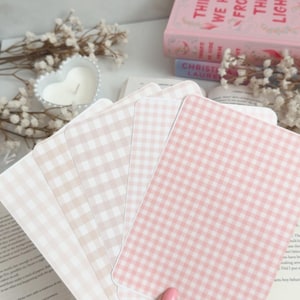 Könnte beinhalten: Ein Stapel Karten mit rosa und beigem Vichy-Muster. Die Karten werden in einer Hand gehalten, mit einem Buch und einer Kerze im Hintergrund. Der Buchtitel lautet "THINGS WE HIDE FROM THE LIGHT".