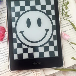 Op de afbeelding: Een zwarte Kindle e-reader met een doorzichtige beschermhoes. Het scherm toont een witte smiley op een zwart-wit geblokt patroon. Het woord "kindle" staat onderaan afgedrukt.