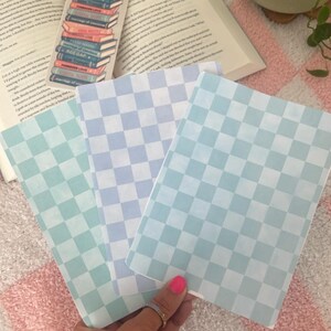 Könnte beinhalten: Drei Notizbücher mit karierten Mustern in Blau- und Grüntönen werden gehalten. Ein Lesezeichen mit einem Bücherstapel ist sichtbar, zusammen mit einem offenen Buch und einer kleinen Topfpflanze. Der Hintergrund ist eine rosa-weiß karierte Decke.