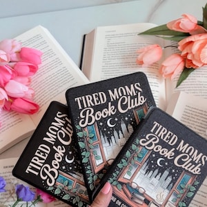 May include: Three black book covers with the text "TIRED MOMS Book Club" and a window design with a cityscape, books, and a coffee cup. The covers are surrounded by open books and pink flowers.