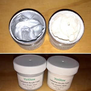 May include: Two small jars of reflective acrylic paint. The jar on the left is silver and the jar on the right is white. Both jars have the label "ViziGlow" and say "Reflective Acrylic Paint". The silver jar says "Ultra-Bright Silver" and the white jar says "High-Visibility White". Both jars have a label that says "3 fluid oz / 90 ml" and "Stir Well!"