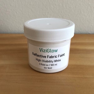 May include: A white jar of ViziGlow Reflective Fabric Paint in High-Visibility White. The jar contains 3 fluid ounces or 180 milliliters of paint. The label instructs to stir well.