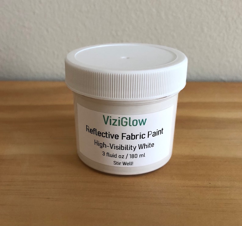 May include: A white jar of ViziGlow Reflective Fabric Paint in High-Visibility White. The jar contains 3 fluid ounces or 180 milliliters of paint. The label instructs to stir well.