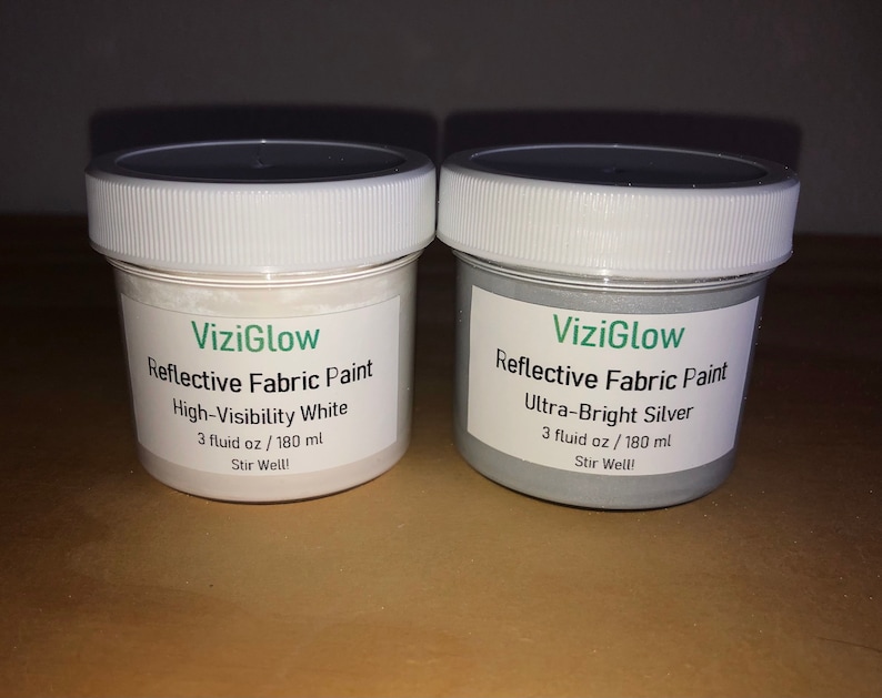 May include: Two small jars of ViziGlow reflective fabric paint. One jar is white and labeled "High-Visibility White". The other jar is silver and labeled "Ultra-Bright Silver". Both jars contain 3 fluid ounces (180 ml) of paint and have the instruction "Stir Well!" printed on the label.