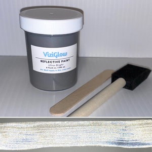 May include: A small grey jar of reflective paint labelled "ViziGlow Reflective Paint Ultra-Bright 8 fluid oz / 240 ml Stir Well! Apply in thin, even coats". The jar is next to a wooden paint stirrer and a foam paint brush.