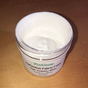 May include: A white jar of ViziGlow Reflective Fabric Paint, labeled "High-Visibility White", with a 3 fluid ounce (180 ml) capacity. The jar is open and the paint is visible inside.