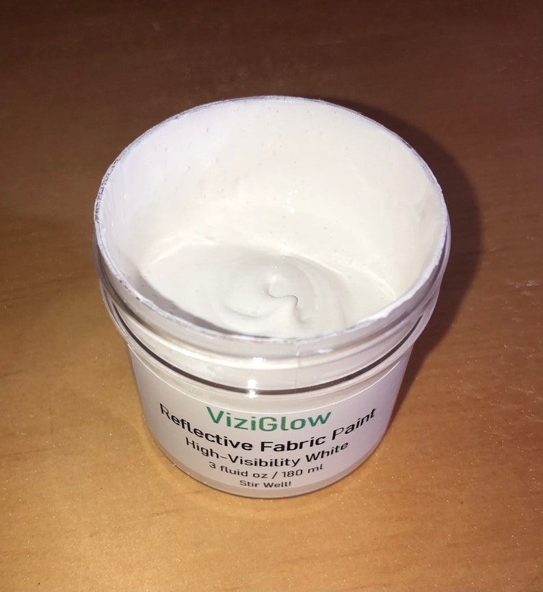 May include: A white jar of ViziGlow Reflective Fabric Paint, labeled "High-Visibility White", with a 3 fluid ounce (180 ml) capacity. The jar is open and the paint is visible inside.