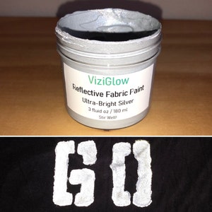 May include: A silver jar of ViziGlow reflective fabric paint. The label reads "Ultra-Bright Silver" and "3 fluid oz / 180 ml". The jar is on a wooden surface. The paint is shown applied to black fabric, creating a reflective "GO" design.