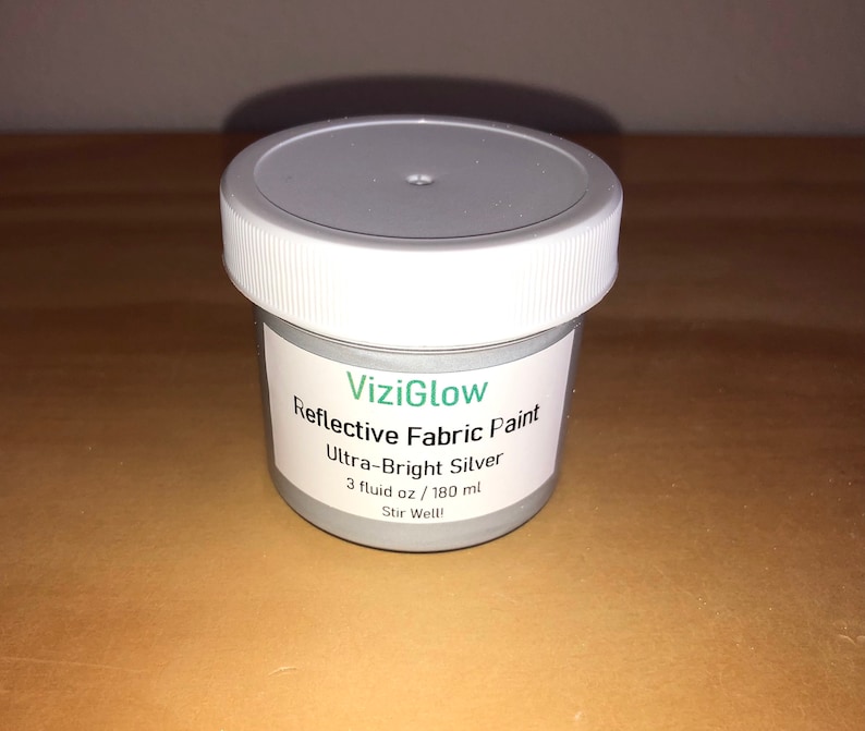 May include: A small, white jar of ViziGlow reflective fabric paint in ultra-bright silver. The jar contains 3 fluid ounces (180 ml) of paint and has a label that says "Stir Well!"