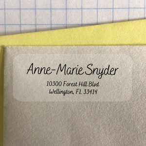 May include: A clear, rectangular address label with rounded corners, featuring the name "Anne-Marie Snyder" and an address in black script. The label is on a light gray surface, with a yellow and white background.