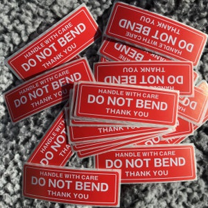 May include: A collection of red rectangular stickers with white text. Each sticker reads "HANDLE WITH CARE, DO NOT BEND, THANK YOU". The stickers are scattered on a textured gray surface.