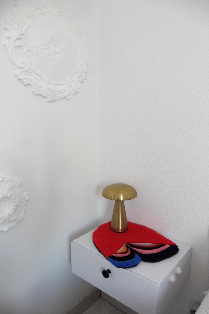 Op de afbeelding: Een goudkleurige paddenstoelvormige lamp staat op een wit nachtkastje. Onder de lamp ligt een rood, veelkleurig tapijt. De witte muur is voorzien van decoratief stucwerk.