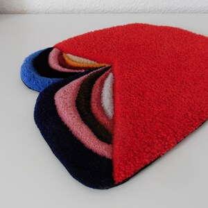 Op de afbeelding: Een abstract tapijt in hartvorm, met een rood gedeelte en een waaier van gebogen delen in blauw, roze, bruin en wit. Het tapijt is gemaakt van een zacht, getextureerd materiaal. Het ontwerp van het tapijt is modern en opvallend.