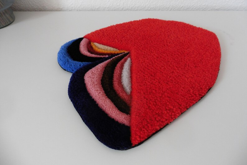 Op de afbeelding: Een abstract tapijt in hartvorm, met een rood gedeelte en een waaier van gebogen delen in blauw, roze, bruin en wit. Het tapijt is gemaakt van een zacht, getextureerd materiaal. Het ontwerp van het tapijt is modern en opvallend.
