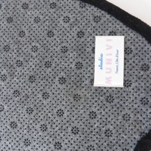 Op de afbeelding: Close-up van een grijze, getextureerde stof met een herhalend bloemenpatroon. Een zwarte rand omlijst de rand. Een wit label met de tekst "studio MUNIVI France, C&ocirc;te d'Azur" is aan de stof bevestigd.