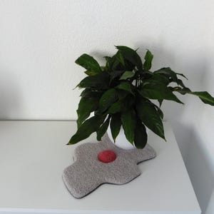 以下が含まれることがあります： 白い鉢植えの観葉植物が、赤い中心部がある灰色の花型のマットの上に置かれています。植物は濃い緑色の葉をしています。これらのアイテムは、白のドレッサーの上にあります。