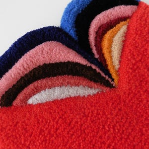 Op de afbeelding: Een kleurrijk, getextureerd tapijt met een regenboogontwerp. Het tapijt heeft lagen van blauwe, roze, bruine, witte en oranje bogen tegen een felrode achtergrond. Het tapijt is gemaakt van een zacht, pluche materiaal.