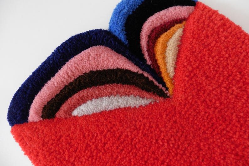 Op de afbeelding: Een kleurrijk, getextureerd tapijt met een regenboogontwerp. Het tapijt heeft lagen van blauwe, roze, bruine, witte en oranje bogen tegen een felrode achtergrond. Het tapijt is gemaakt van een zacht, pluche materiaal.