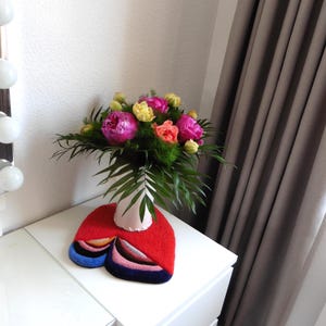 Op de afbeelding: Een kleurrijk boeket van roze, gele en oranje bloemen in een witte vaas op een witte tafel. Onder de vaas ligt een rood vilten tapijt met een abstract ontwerp en blauwe, roze en zwarte accenten. Grijze gordijnen en een witte muur op de achtergrond.