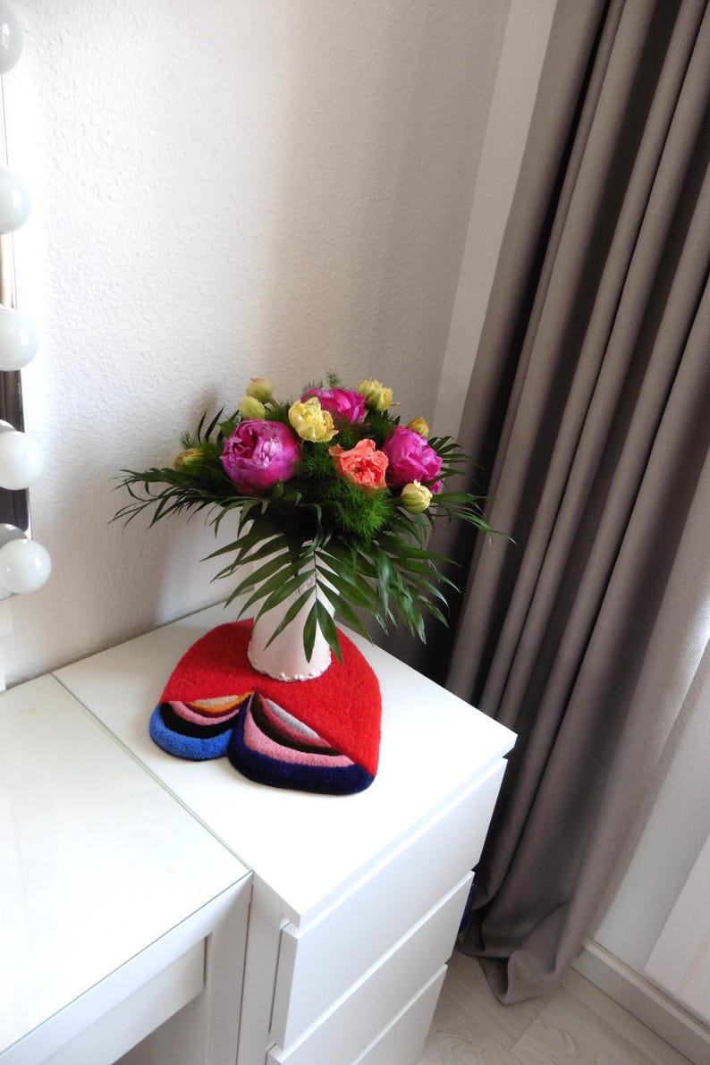 Op de afbeelding: Een kleurrijk boeket van roze, gele en oranje bloemen in een witte vaas op een witte tafel. Onder de vaas ligt een rood vilten tapijt met een abstract ontwerp en blauwe, roze en zwarte accenten. Grijze gordijnen en een witte muur op de achtergrond.