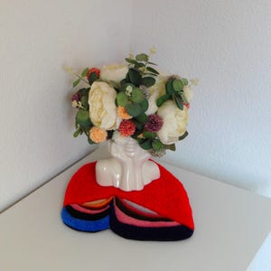 Op de afbeelding: Een witte keramische bustevase met een bloemstuk van cr&egrave;mekleurige pioenrozen, oranje pompons en groen blad. De vaas staat op een rode vilten mat met kleurrijke strepen in blauw, roze en zwart.