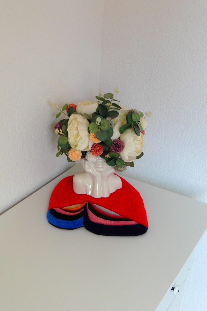 Op de afbeelding: Een witte keramische bustevase met een bloemstuk van cr&egrave;mekleurige pioenrozen, oranje pompons en groen blad. De vaas staat op een rode vilten mat met kleurrijke strepen in blauw, roze en zwart.