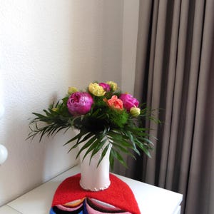 Op de afbeelding: Een boeket van roze pioenrozen, gele tulpen en oranje bloemen in een witte vaas, rustend op een rode vilten mat met blauwe, roze en bruine accenten. De opstelling staat op een witte tafel.