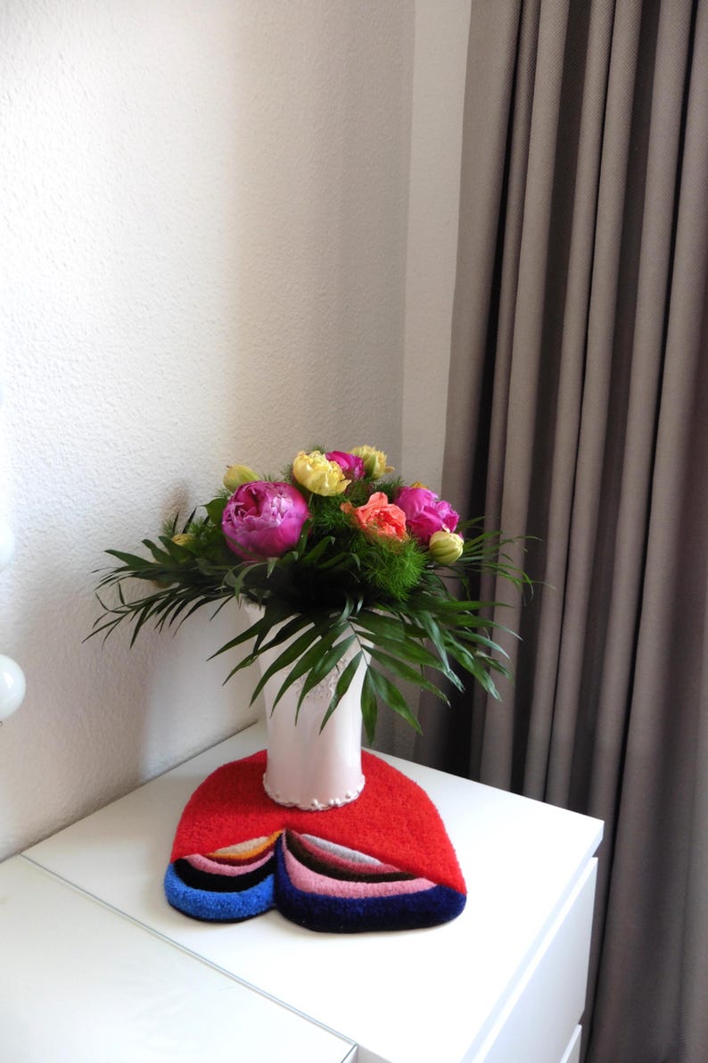 Op de afbeelding: Een boeket van roze pioenrozen, gele tulpen en oranje bloemen in een witte vaas, rustend op een rode vilten mat met blauwe, roze en bruine accenten. De opstelling staat op een witte tafel.
