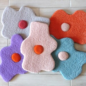 以下が含まれることがあります： グレー、紫、ピンク、オレンジ、青の色合いの、5つのカラフルな花の形をしたラグ。各ラグの中央には、対照的な円形のアクセントがあります。ラグは、テクスチャ加工された素材で作られています。